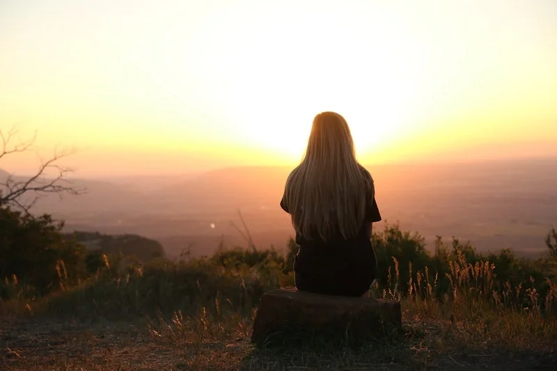 Girl sitting on a tree trunk back to camera facing sunset. Dealing with your mortality